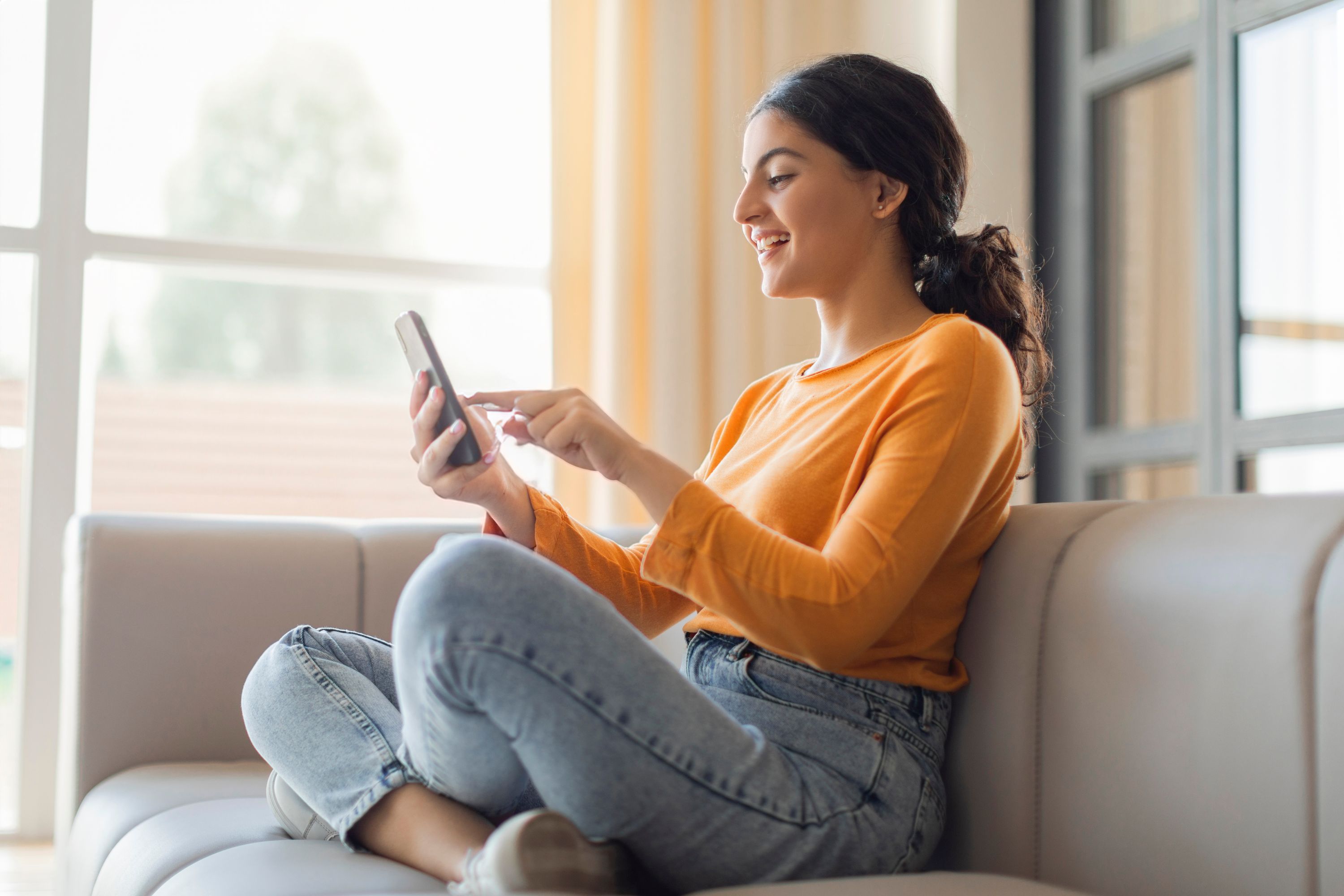 Frau Auf Couch im schneidersitz orangener Pulli, bedient Ihr handy